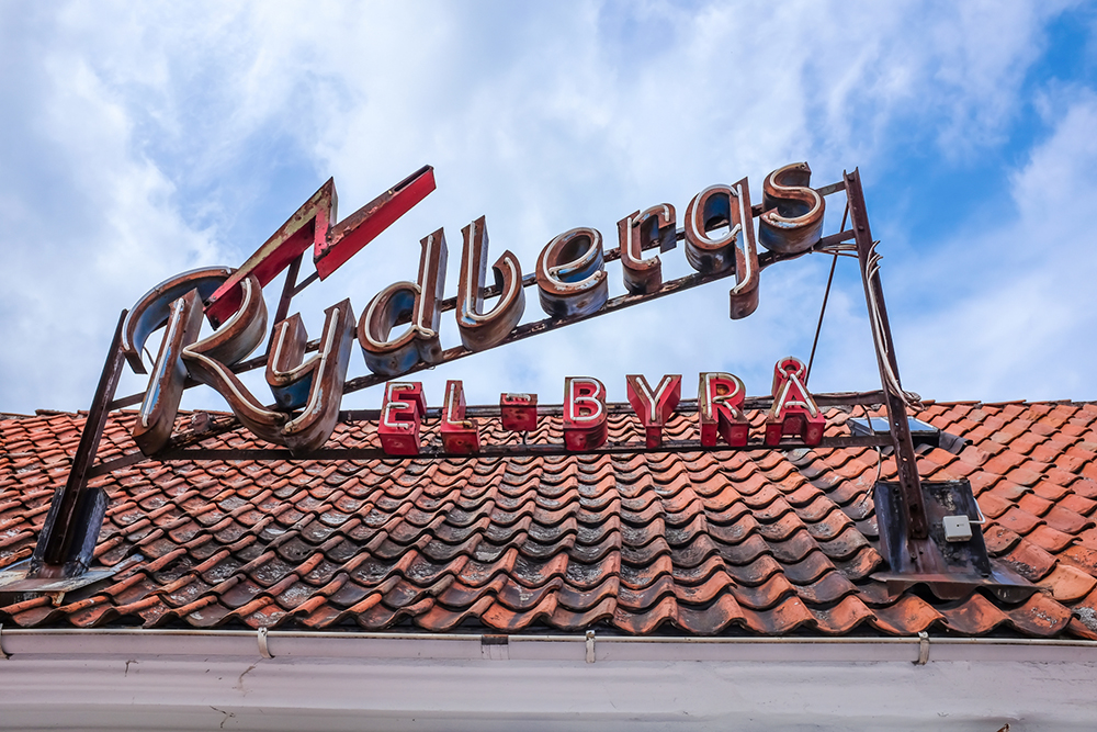 Fotografi föreställande en äldre neonskylt med texten Rydbergs el - byrå som sitter ovanpå ett tak.