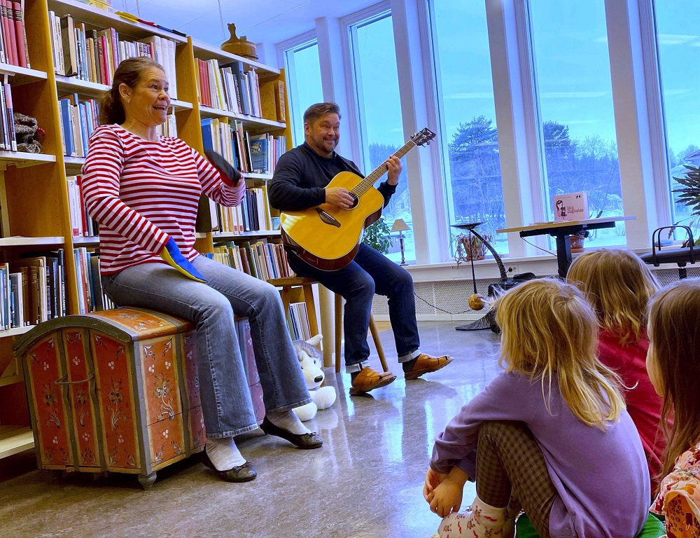 En kvinna sjunger och en man spelar gitarr för en grupp barn i ett bibliotek. 