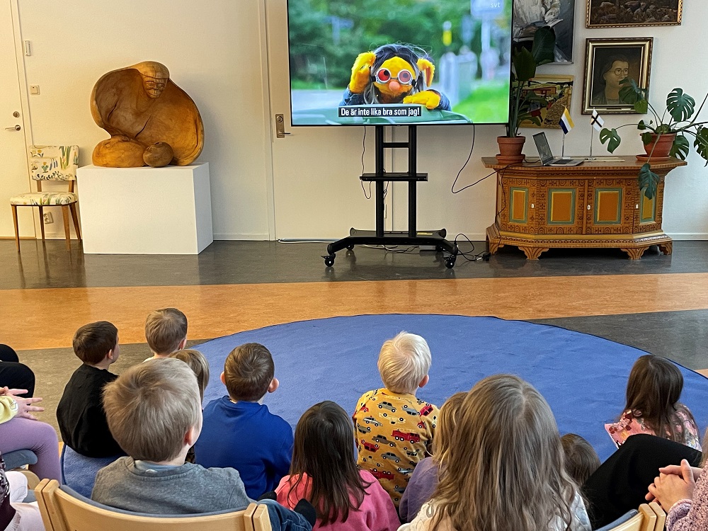 Många barn sitter och tittar på en tv där ett barnprogram visas.