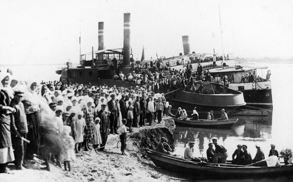 Svartvitt fotografi med hundratals människor som står uppställda framför två ångfartyg som ligger vid strandkanten.